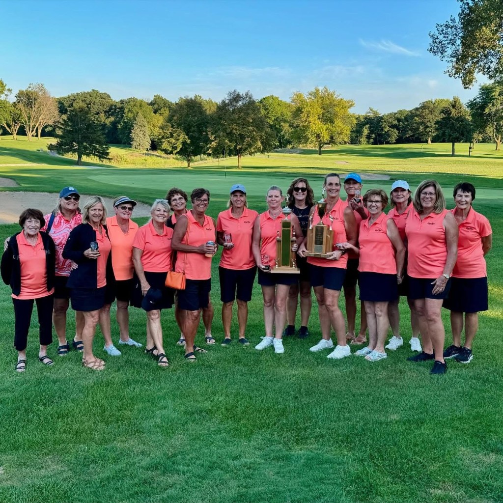A large group of ladies all wearing the same pinkish shirt smile for the camera. The 2 near the center are holding trophies.