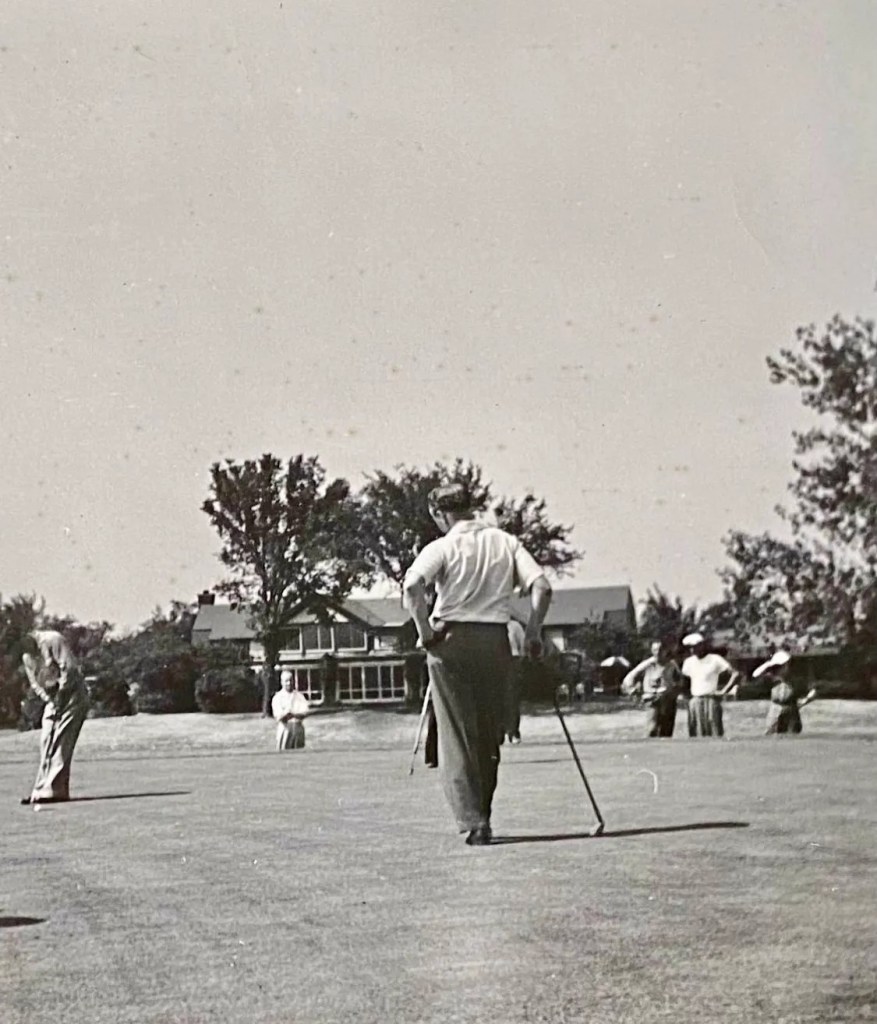 A black and white photo of a gentleman standing off to the side as another gentleman preps his swing.