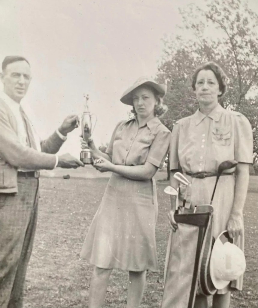 A black and white photo of a gentleman holding a trophy with a woman. Another woman stands off to the side.